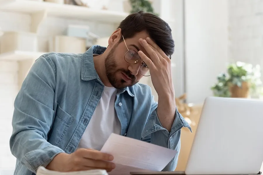 A man with his hand on his forehead holding a foreclosure document by a laptop