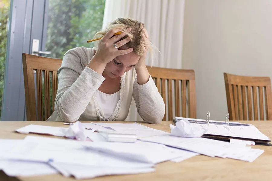 A woman sitting at a table surrounded by bills holding both her hands to her head