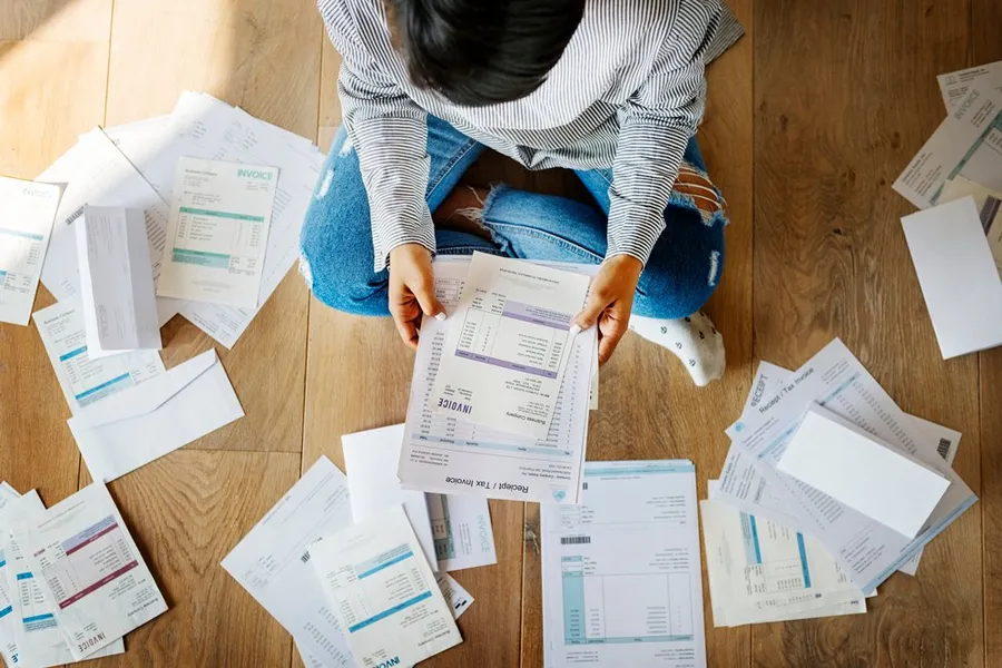 Person sitting on floor with scattered bills, holding one up.