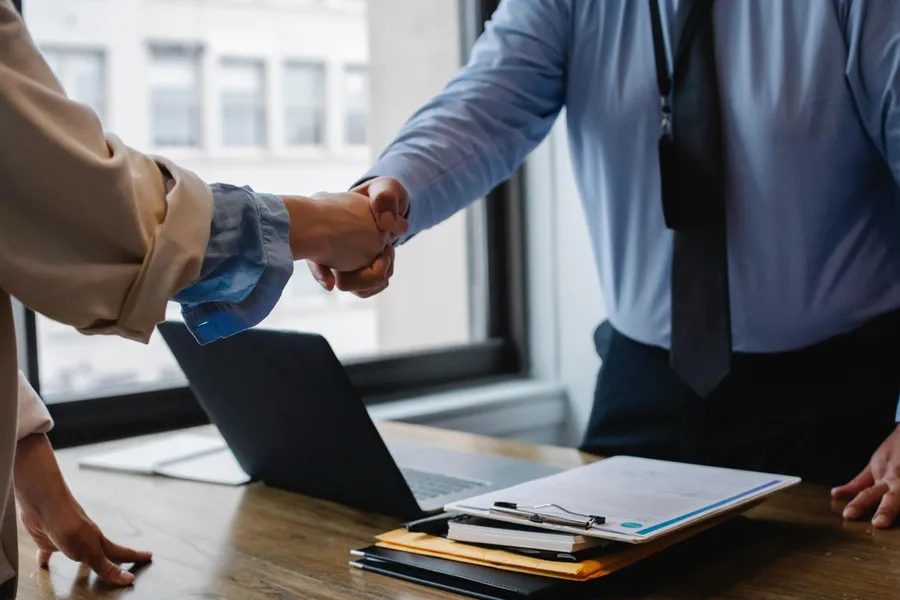 An attorney and client shaking hands over a desk with a laptop and documents.