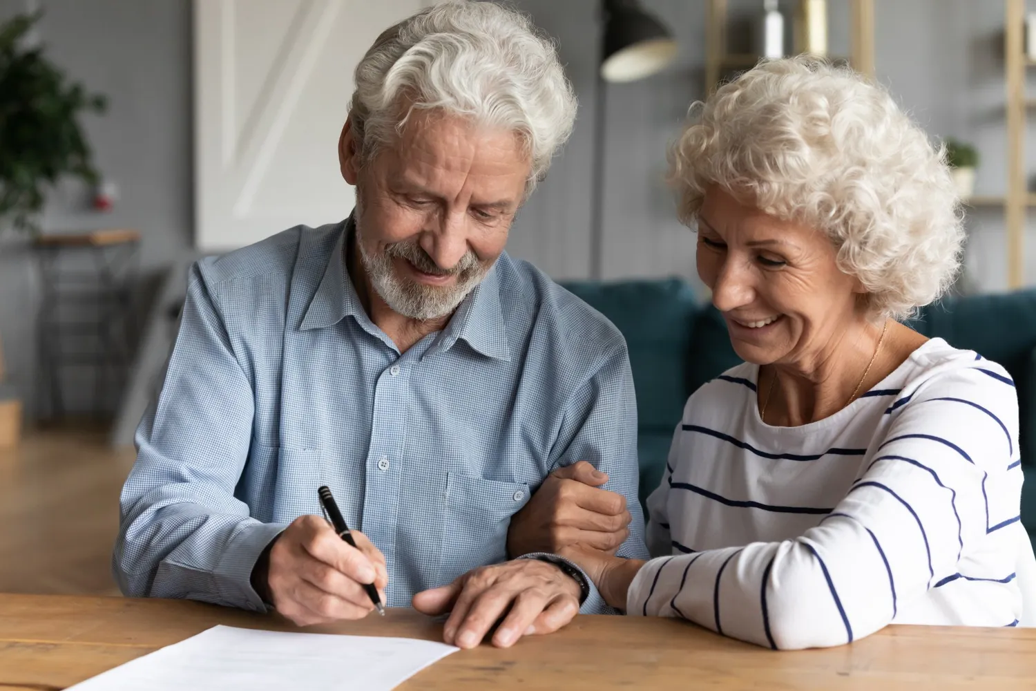 Two elderly individuals sitting at a table, one signing a will.