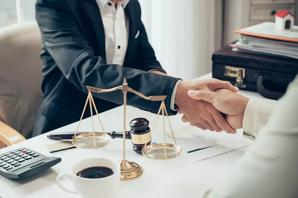 Attorney sitting at desk filled with a court gavel, Scales of Justice, calculator, and notebooks shaking a man's hand